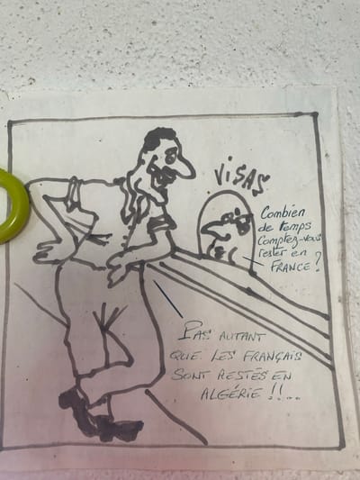 A hand-drawn cartoon, rendered in dark ink on a light-colored sheet of paper, is affixed to a white, textured wall. A circular, light-green clip secures the paper on its left edge. The cartoon illustrates two stylized figures. On the left, a male figure with dark hair, a prominent nose, and an expressive face leans forward, resting his hands on a counter or barrier. On the right, a simpler head, representing an official, is visible above the counter. The word "VISAS" is written above the official's head. French text to the right of the official reads: "Combien de temps comptez-vous rester en FRANCE?" (How long do you intend to stay in FRANCE?). Below the counter, the response text states: "PAS AUTANT QUE LES FRANÇAIS SONT RESTÉS EN ALGÉRIE !!" (Not as long as the French stayed in ALGERIA!!). This scene is located in the city of La Salle-les-Alpes, France.