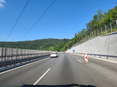 This image captures a view from a vehicle driving on a multi-lane highway through a mountainous, forested area. The highway is paved with asphalt and marked with white lines. On the left side of the road, there is a metal guardrail and a sound barrier made of metal mesh and concrete panels. On the right side, a grey concrete retaining wall slopes upwards, topped with a metal fence. Trees and dense green foliage cover the hillsides in the background. Several vehicles are visible on the highway, including a white car in the foreground and another car further ahead. Traffic cones, including a distinctive orange and white striped marker and a blue directional sign, are present on the right side of the road, suggesting a lane closure or construction. Overhead, power lines stretch across the clear blue sky. The scene appears to be on the Hemus Motorway (A2) near Vrachesh, Bulgaria, a route known for its scenic mountainous terrain.