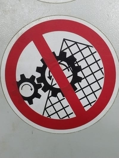 A circular red prohibition sign with a red diagonal line is overlaid on a white background. Inside the circle, two black gears are depicted, with arrows indicating rotation. Behind the gears, a black grid pattern is visible, resembling a mesh or fence. The sign is affixed to a textured, light gray surface, possibly a wall or machine casing. The image is a close-up of the sign, with the surrounding environment out of focus. The location is stated as Гожскі сельскі Савет, Belarus.
