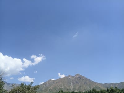 The image shows a mountain range under a clear blue sky with scattered clouds. In the foreground, there are trees and foliage, suggesting a natural landscape. The mountains are rugged with visible slopes and some sparse vegetation. The sky occupies a significant portion of the image. There are no people or discernible actions visible. The environment appears to be rural or mountainous, likely in or around the city of Kalaynay, Tajikistan, based on the provided context.