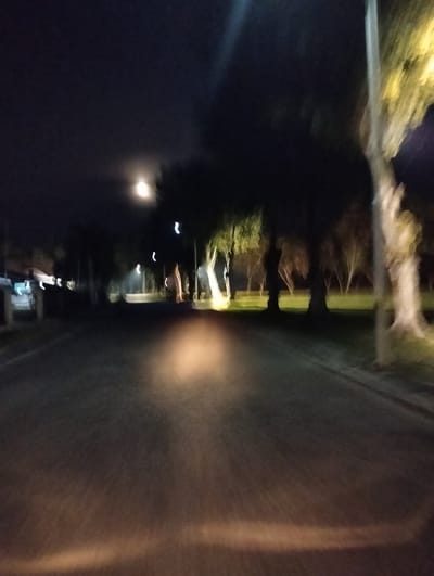 This media file depicts a nighttime scene, likely taken from a moving vehicle, on a road bordered by trees. The image is blurry due to motion and low light conditions, characteristic of a night photograph. The subjects are primarily the road itself, illuminated by car headlights in the foreground, and a line of trees on either side of the road. There are also some indistinct structures visible on the left side of the road, possibly houses or buildings.
The activity appears to be travel, with the perspective suggesting the viewer is a passenger or driver. The time of day is clearly night, with a visible moon in the upper left portion of the frame, partially obscured by clouds. The weather appears to be clear enough for the moon to be visible, though some atmospheric haze or fog might be contributing to the blurriness.
Notable details include the soft glow of streetlights or house lights in the distance, creating streaks of light typical of long exposure photography in motion. The trees on the right side are particularly prominent, with their trunks and branches visible against the dark sky. The overall mood is somewhat serene and perhaps a bit mysterious, given the nighttime setting and the blurred visuals. There is no visible text in the image. The location context provided, Palohgadeng, Indonesia, suggests a rural or suburban setting where such a tree-lined road at night would be common.