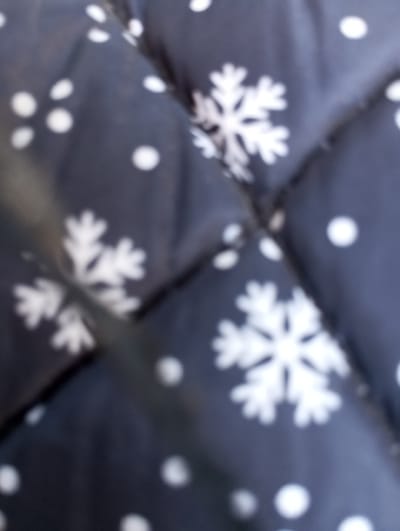 The image shows a close-up, blurry view of a dark blue fabric with a pattern of white snowflakes and polka dots. The fabric appears to be quilted, with visible stitching lines creating a grid-like texture. The lighting is soft and diffused, and the focus is not sharp, giving the image an impressionistic quality. There are no people, animals, or identifiable landmarks from the city of Voznesenske, Ukraine, visible in the frame. The scene is solely focused on the textile pattern.