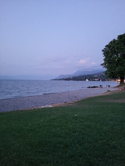 A serene waterscape captures a lake bordered by a pebble beach and lush green grass. Distant mountains fade into the evening sky, mirroring a tranquil twilight ambiance. A tree's leafy canopy extends into the upper left corner, while faint city lights punctuate the horizon. Several people are visible along the beach.