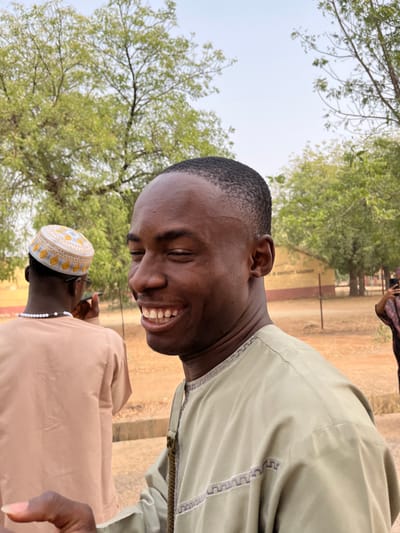 This image depicts three individuals outdoors in Kanti, Nigeria. The primary subject, a Black male in his 20s, is positioned in the foreground, facing right but looking towards the camera with a broad, open-mouthed smile revealing his teeth. He has a short, faded haircut and wears a light olive-green long-sleeved garment with grey and white geometric embroidery on the chest and sleeves. His left hand is partially visible in the lower left.
Behind the primary subject, to the left, a second Black male is seen from the back, wearing a light brown long-sleeved garment and a white kufi cap with yellow and grey embroidered patterns. A white beaded necklace is visible around his neck. He holds a dark-colored smartphone to his right ear. A third individual is partially visible on the far right, also appearing to hold a smartphone.
The ground is dry, reddish-brown earth. In the background, several trees with green foliage and visible branches are present, along with a low-rise building featuring faded reddish-orange walls. A faint, dark inscription on a lighter background is visible on the building, suggesting a sign, possibly indicating a school or institution. The sky above is a clear, light blue.