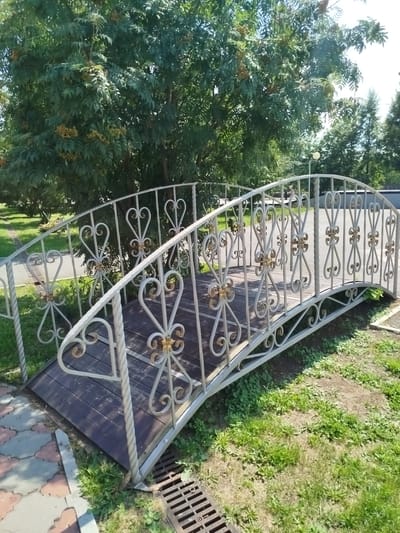 The image shows a decorative arched metal bridge with ornate railings, made of white metal with gold accents. The bridge has a dark wooden deck. It is situated outdoors, in a park-like setting. Lush green trees with some berry clusters are visible in the background. The ground around the bridge is covered with grass and some paving stones are visible on the left side, leading to the bridge. A metal grate, possibly a drain cover, is visible near the base of the bridge on the right. The scene appears to be captured on a sunny day. The bridge is located in the city of Yalutorovsk, Russia.