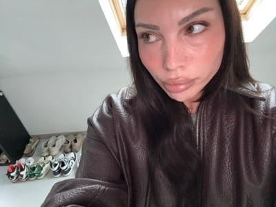 The image depicts a female individual in an indoor setting, likely an attic room, characterized by a white sloped ceiling and a wooden-framed skylight in the upper right. The individual, positioned in the foreground, has dark, long hair, light freckles visible on her nose, and wears noticeable lip gloss. She is clad in a dark brown, textured, zippered jacket over a light grey garment, with her gaze directed towards the upper right of the frame. In the background, along the lower left wall, approximately eleven pairs of casual shoes, including sneakers in various colors, are arranged in two rows on a light-colored floor. A dark, angular object is partially visible leaning against the wall on the far left. The specific location within Tielrode, Belgium, cannot be determined from the visual information presented in the image.