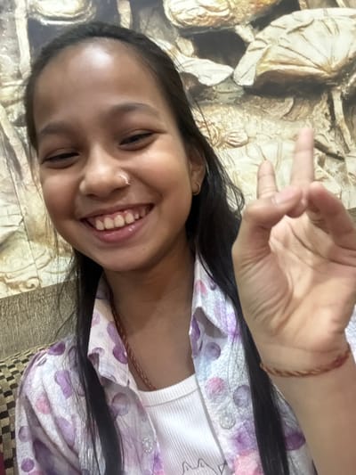 A smiling young woman poses with her head slightly tilted, showing her face clearly. She wears a white shirt with a floral, patterned lavender shirt on top. Her black hair is smooth and shiny. She is wearing a nose ring and earrings, with a bracelet on her wrist. Her hand is up, displaying the peace sign. The background is a mix of textured beige and gold with some patterned fabric.