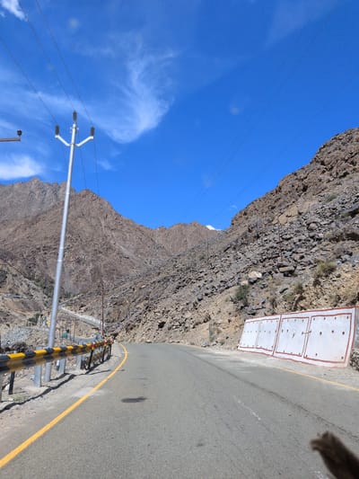 A paved road curves through a mountainous landscape under a bright blue sky with scattered white clouds. The road is bordered by a yellow and black striped guardrail on the left. Steep, rocky hillsides rise on either side of the road, sparsely covered with dry vegetation. Power lines are visible strung between utility poles that ascend the slopes. In the foreground on the right, there are several white panels with red trim, likely part of a retaining wall or drainage system. The scene appears to be an isolated, high-altitude area, possibly in the city of Chanigund, India, given the context.