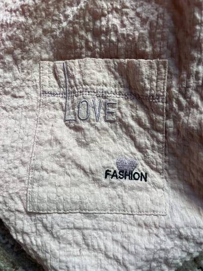 The image is a close-up shot of a light pink, textured fabric, likely a garment. On the fabric, there are two embroidered phrases. On the left, the word "LOVE" is embroidered vertically in a light purple thread, with each letter stacked atop the other. To the right of "LOVE," and slightly higher, the word "FASHION" is embroidered vertically in black thread, with a small, stylized heart shape preceding it. The fabric itself has a crinkled texture. The lighting suggests a bright, possibly daytime setting, casting shadows that highlight the texture of the material. The image focuses solely on the detail of the embroidery and fabric, with no broader scene or subjects visible. The location context of Ташлак, Kyrgyzstan, is provided, but no visual cues within the image itself confirm this location.
