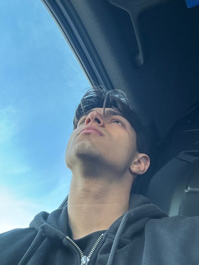 An individual is depicted from the chest up, situated within the interior of a vehicle in Kempten (Allgäu), Germany. The person, a male with dark hair, is wearing a dark, zippered hooded sweatshirt. His head is tilted backward, and his gaze is directed upwards towards an open sunroof or moonroof. Through this opening, a clear blue sky with minimal white cloud cover is visible, indicating daylight conditions. The dark interior trim of the vehicle's ceiling and sunroof frame occupies the upper right portion of the image. No other individuals or distinguishing features of the surrounding city are visible.