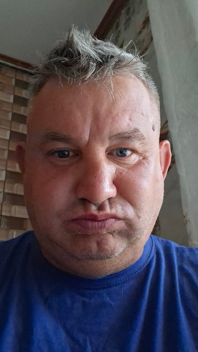 A close-up, head-and-shoulders selfie taken by a fair-skinned, middle-aged Caucasian male with short, spiky grey hair and blue eyes. He is wearing a bright blue t-shirt. His cheeks are puffed out, and his lips are pursed, suggesting he is holding his breath or making a funny face. The background is blurred, but appears to be an indoor setting with textured walls and a curtain on the right. There is a dark mole on the man's forehead. The image is taken at a slightly upward angle, with the camera positioned in front of the man. The lighting is natural, coming from the front and slightly from the right. The overall impression is a candid, informal portrait. No specific landmarks or references to Voinova, Moldova are visible in the image.
