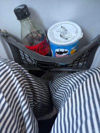 In a seat-back pocket on what appears to be public transport, a Coca-Cola bottle with a black cap and a Pringles can are placed. The Coca-Cola bottle is partially filled with a dark liquid and has a red label. The Pringles can is blue with the Pringles logo featuring a caricature of a man with a mustache. The seat-back pocket is made of gray mesh material. In the foreground, a person's lap is visible, covered by striped pants featuring alternating navy blue and white stripes. The background is a plain, light-colored surface, possibly the back of a seat. The image does not contain any information or visual cues to identify its specific location in the city of Pombia, Italy.