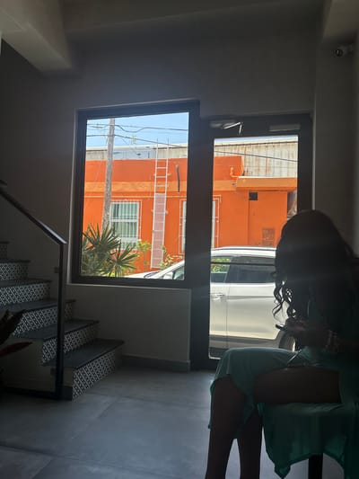 An over-the-shoulder view shows a woman with dark, curly hair sitting in a chair indoors, facing a large window and a glass door. She is silhouetted against the bright outdoor light, holding a smartphone in her right hand. She wears a light teal dress. To her left, a staircase with black and white patterned risers ascends. Through the window and door, an orange building is visible across the street, with a white ladder attached to its side. A white car is parked in front of the building. The floor inside is tiled in large grey squares. The scene is likely in San Juan, Puerto Rico, given the architectural style and vibrant colors common in the city.