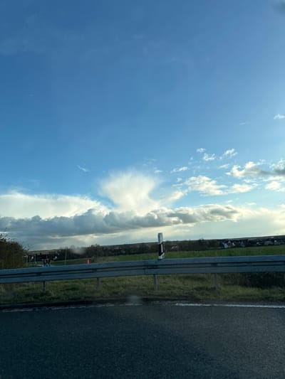 The image depicts a view from a moving vehicle, likely on a highway or roadway in Herzogenaurach, Germany. On the right side of the frame is a section of asphalt road and a roadside barrier. Beyond the barrier is a grassy area bordering a residential area, with houses visible in the background. The left side of the image is dominated by a sky filled with a mix of cirrus and cumulus clouds. The overall scene suggests a slightly overcast day.