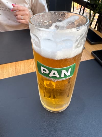 The image shows a close-up of a glass of beer with a label reading "PAN". The beer is golden yellow and topped with a frothy head. The glass is partially filled and condensation is visible on its exterior. The glass rests on a dark textured surface, likely a table. In the background, a person's arm and hand, adorned with a ring and bright pink nail polish, are visible, resting on a beige garment that has some embellishments. The setting appears to be outdoors, with sunlight casting reflections on the glass and a glimpse of a railing and greenery in the background, suggesting a café or restaurant environment, possibly in Makarska, Croatia.