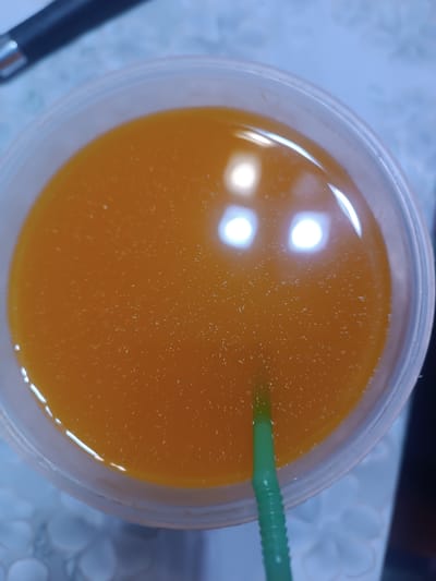 The image shows a close-up, overhead view of a clear plastic cup filled with a bright orange liquid, likely juice. A green plastic straw is partially submerged in the liquid. The surface of the liquid is slightly disturbed, with small bubbles or particles visible. Two bright, circular reflections are visible on the surface of the liquid, suggesting overhead lighting. The cup is transparent, revealing the liquid within. The background is out of focus but appears to be a patterned surface, possibly a tablecloth or placemat, with hints of blue and white. To the upper left of the cup, a dark object with a metallic tip is partially visible, possibly a pen or stylus. The overall impression is of a refreshing beverage on a table. There is no discernible connection to the city of Бидайкөл, Kazakhstan in the visual information provided by the image.
