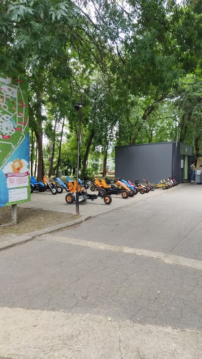 The image captures a row of pedal go-karts lined up on a paved walkway in a park setting, likely in the city of Novaya Adygeya, Russia, as indicated by the visible map sign with Cyrillic text that reads "ПАРК ПОБЕДЫ" (Victory Park). The go-karts are brightly colored, primarily orange, blue, yellow, and pink, and appear to be designed for children or recreational use. Some of the go-karts have double seats.
In the background, there is a modern, dark gray building with a green trim, possibly a rental facility or information center, with a few people standing near it. The area is surrounded by lush green trees, suggesting a pleasant outdoor environment. A lamp post stands near the center of the go-kart display. The ground is paved with interlocking concrete blocks, transitioning to asphalt on the foreground pathway. A tall, narrow sign with a map and text is visible on the left side of the image. The sky is overcast, indicating a cloudy day.