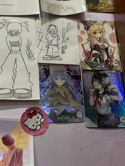 The image displays a collection of anime-style trading cards and drawings arranged on a flat surface, likely a desk or table. In the upper left corner, text in Russian is partially visible. To the left of the trading cards, two hand-drawn sketches of anime characters are present. The trading cards themselves feature colorful illustrations of female anime characters. A pink Hello Kitty sticker is adhered to the surface in the lower left portion of the image. In the lower left of the image, a portion of a poster or print is visible, showing the back of a person looking upwards. The overall scene appears to be a personal collection of artwork and merchandise. There is no discernible indication of a specific location within a city, including ЖК «Борисоглебское», Russia, or any specific urban context. The image focuses solely on the displayed items.