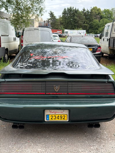 The image captures the rear view of a dark green Pontiac Firebird parked outdoors in a gravel lot, likely in the vicinity of Reckange-sur-Mess, Luxembourg, given the presence of a European license plate. The car has a prominent spoiler and a distinctive red stripe across its taillight bar. A California license plate with the numbers "23492" is mounted on the rear bumper. Two exhaust pipes are visible below the bumper. The car's rear window has red decals and a smaller white sticker. In the background, several vans, including white panel vans, and other vehicles are parked. A line of trees and a cloudy sky form the backdrop. The environment appears to be a commercial or industrial area, possibly a parking lot for businesses or vehicles.