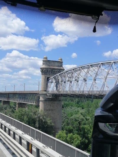 The image shows a view of the Cernavoda Bridge in Cernavoda, Romania, from a vehicle. The bridge is a large, steel truss bridge with stone pillars. It spans a body of water, likely the Danube River, which is not visible in the frame. The bridge is surrounded by lush green trees. The sky is blue with fluffy white clouds. The perspective suggests the viewer is looking out from a car or bus, with part of the vehicle's interior, including a side mirror, visible in the foreground. There are no people visible in the image. The bridge appears to be functional, with overhead electrical lines suggesting it is used by trains.