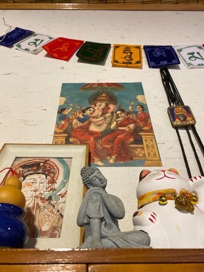 This image shows a wall adorned with various decorative items. Prominently featured is a painting depicting a Hindu deity with multiple arms, surrounded by figures in traditional Indian attire. To the right of the painting, a white cat figurine, possibly a Maneki-neko, is positioned on its side, decorated with gold bells and a yellow and orange sash. Below the cat figurine and to the right of the Hindu painting, a framed artwork displays a portrait of a person with ornate details. A gray, Buddha-like statue is resting above the framed artwork.

Hanging on the wall to the left of the Hindu painting is a string of small, colorful pennants. These pennants are blue, yellow, green, and red, and each appears to have symbols or text printed on them. Some of these symbols resemble Tibetan script. A thin black cord with a small, square pendant featuring a portrait is also strung horizontally across the wall above the Hindu painting. The wall itself has a textured, off-white surface, and the overall scene suggests an indoor setting, likely a home or a shop, given the collection of decorative elements. The lighting appears to be artificial, casting soft shadows. The arrangement of objects indicates a cultural or spiritual theme. There is no discernible information about the time of day, weather, or specific emotions conveyed by the subjects. The location context provided is Dujiangyan City, China.