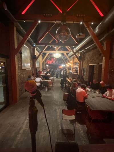 This image depicts the interior of a spacious, high-ceilinged event venue, likely a converted industrial building or barn, located in Marquette-lez-Lille, France.
The scene is dimly lit with warm ambient light, complemented by distinctive red LED accent lighting integrated into the dark wooden ceiling trusses and structural beams. A large, reflective disco ball hangs centrally from the ceiling, alongside an ornate chandelier further in the background. Exposed red brick walls define the periphery of the long, rectangular room. Ventilation ducts run along the upper right wall and ceiling.
In the immediate lower-left foreground, a professional-grade microphone with a pink-orange foam windscreen is positioned on a dark stand. An empty, light-colored chair with a metal frame sits to the right of the microphone stand. The floor is dark and appears to be made of wood.
The mid-ground and background are populated by an estimated 30 to 50 adults. Various seating arrangements include smaller, square tables on the left, occupied by groups of individuals, and a long, dark communal table on the right where approximately seven individuals are seated, some engaged in conversation, others observing mobile devices. Drinks are visible on the tables. Further back, a bar area with bottles and glassware is discernible on the right side. At the very rear of the venue, an elevated section displays strong red backlighting illuminating indistinct red shapes or artwork. Patrons throughout the venue are casually dressed and appear to be engaged in social interaction, dining, and drinking. A large, light-toned abstract artwork is mounted on the exposed brick wall on the left side of the room.