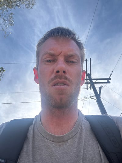 An adult male subject is depicted in an upward-angled selfie, looking directly at the camera. He has fair skin, short light-colored hair, light blue eyes, and a short beard/stubble. His brow is furrowed, and his lips are slightly parted. He is wearing a light gray crew-neck t-shirt and a black backpack, with the "OGIO" brand logo visible on the left shoulder strap. The background consists primarily of a bright blue sky with wispy clouds. In the upper right quadrant, a utility pole with multiple cross-arms and associated overhead power or communication lines is visible. Additional overhead lines extend across the upper center of the frame. Sparse green tree foliage is visible in the upper left corner. The lighting suggests a bright, sunny daytime environment. The scene is located in the city of Long Beach, United States of America.