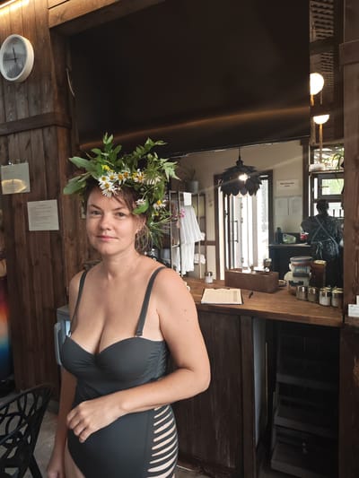A woman wearing a dark grey one-piece swimsuit and a flower crown stands in front of a wooden counter. The swimsuit has cut-out details on the sides. She is looking directly at the camera with a neutral expression. Behind her, a person wearing an apron is visible working at a counter. The environment appears to be an indoor establishment with wooden walls and a rustic aesthetic. Various items are on the counter, including containers, bottles, and a clipboard with papers. A clock with a black frame and a nature-themed face is mounted on a wooden wall to the left of the woman.