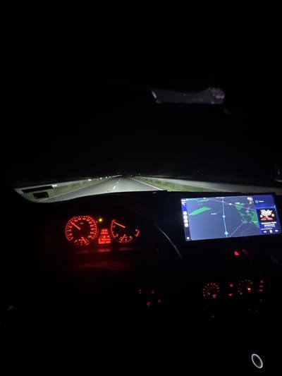 The image depicts the interior of a car at night, likely taken from the driver's perspective. The dashboard and instrument cluster are illuminated in red, displaying speed, RPM, and other vehicle information. A navigation system screen shows a map, indicating the vehicle's location on a road. The road ahead is visible through the windshield, illuminated by the car's headlights.
Given the OCR text "+19.0 D A 10.4 329052 416.1", the coordinates 329052 and 416.1 don't seem to correspond to standard latitude and longitude formats, nor do they immediately suggest a location in Mogila, Bulgaria or elsewhere. The text may represent internal vehicle metrics or non-geographical data.