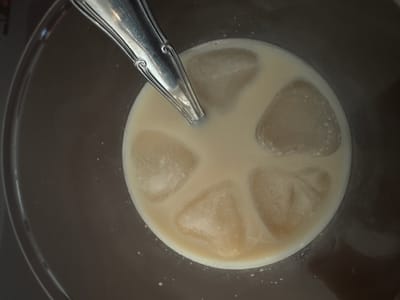 A top-down, close-up shot captures a cold, light brown or creamy beverage in a clear glass. Several irregularly shaped ice cubes float in the drink, some appearing frosty or partially melted. A silver-colored spoon with an ornate handle is partially submerged in the liquid, positioned towards the top center of the frame. The spoon's design features decorative swirls and lines along its handle. The glass rim is visible along the left and bottom edges, suggesting a transparent container. The background is dark and out of focus, providing no specific details about the setting beyond suggesting an indoor environment, possibly a table or counter. No text is visible in the image. The scene suggests a moment of preparing or enjoying a chilled drink.