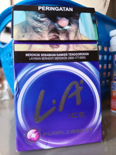 The image shows a pack of LA ICE cigarettes, a brand manufactured by PT. Djarum. The pack features a warning label with an image of a person's throat and mouth, indicating health risks associated with smoking. The front of the pack displays the LA ICE logo in large, stylized letters, with the word "ICE" below it. At the bottom of the pack, there's a circular graphic depicting a berry flavor capsule and the text "PURPLEBOOST." The background suggests the cigarettes are placed on a surface, possibly a table, with a blue basket visible. The presence of "PERINGATAN" (warning in Indonesian) and the cigarette brand itself are strong indicators that this image was taken in Indonesia. Given the specific branding and packaging, this product is commonly sold in convenience stores or kiosks across Indonesia, including areas like Bireuen Meunasah Dayah, which is a locality within the Bireuen Regency of Aceh, Indonesia. The image is a close-up, focusing on the cigarette pack and its details.