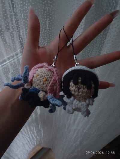The image shows a hand holding two crocheted charms. The charm on the left has pink hair, yellow eyes, and blue appendages. The charm on the right has black hair and purple eyes, with a white fluffy outer layer. Both charms are attached to black strings with silver clasps. The background appears to be a textured white surface, possibly sheer fabric. A timestamp in the lower right corner reads "29.06.2026 19:55". There is no indication of the city of Tyrnyauz, Russia.