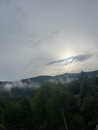 The original image, rotated 90 degrees counter-clockwise, depicts a serene, mountainous landscape under a cloudy sky. The sun, a bright circle of light, is partially obscured by a long, horizontal cloud formation stretching across the upper left quarter of the frame, creating a luminous glare. Dense, dark green forests cover a range of mountains that dominate the right side of the image, extending into the background. Pockets of mist or low-lying clouds are visible clinging to the slopes and bases of these forested mountains, particularly in the lower right and center-right sections, adding an atmospheric quality.

In the immediate foreground on the right, dark tree canopies are prominent. Several thin, dark lines, likely power cables or wires, stretch diagonally across this forested area. A lighter-colored, possibly fallen or dead, tree trunk lies horizontally amidst the dense greenery on the right. Further within the trees, a faint, small structure, possibly a building or cabin, can be glimpsed.

No people or explicit activities are visible. The scene evokes a sense of calm and natural beauty. The combination of the partially visible sun, clouds, and mist suggests either an early morning or late afternoon setting. No visible text is present. The landscape is consistent with a mountainous and forested region like Rudartsi, Bulgaria.