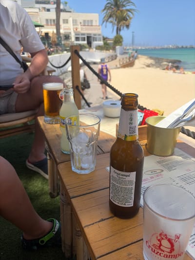 This media file captures a sunny, relaxed scene at an outdoor establishment in La Oliva, Spain, likely a beachside bar or restaurant. The image is taken from a low perspective, looking across a wooden table towards a sandy beach and the turquoise sea under a clear blue sky.

In the foreground, a wooden table holds several items. A half-empty bottle of "Cruzcampo Radler" beer stands prominently, showing text like "ALCOHOL Y SALUD" (Alcohol and Health), "VOL 330 ml e ALC. 2.0% VOL.", and a QR code on its label, along with instructions for consumption and information about the product. Next to it, an empty glass with ice cubes suggests a cold drink has been or is about to be enjoyed. Further on the table, another partially filled beer glass and a small bottle of what appears to be "FANTA" soda are visible. A white plastic "Cruzcampo" branded glass, with a red logo featuring two figures, is also present. A metallic bucket holding cutlery and napkins sits on the table, along with a laminated menu.

Visible menu text includes headings like "All day", "NIBBLES, AND STARTERS", "breads with wick and butter.", and categories such as "FISH" with specific dishes like "SAHAKAN Swa" and "FRESH FISH FROM PUB".

Beyond the table, a person's legs and feet are visible; one foot wears a black and green "CRAFT" branded Croc-style sandal. In the background, palm trees sway, and white buildings line the coast. People are scattered along the sandy beach and near the water, enjoying the bright weather. The overall impression is one of leisure and vacation.