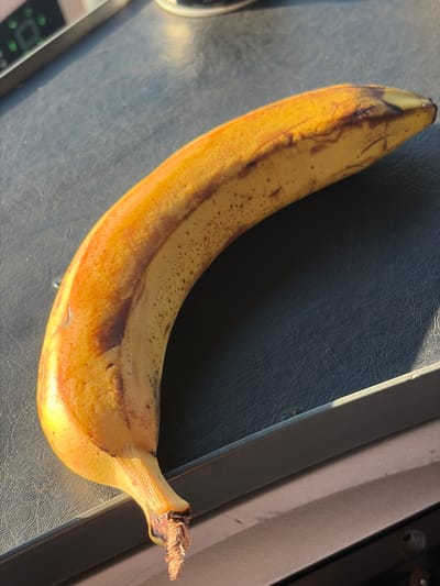 A ripe banana is positioned diagonally on a dark, textured surface, likely a desk or table. The banana is primarily yellow with brown streaks and speckles, indicating its ripeness. The stem end of the banana is visible at the bottom left of the frame and appears somewhat dry and frayed. The lighting suggests it is daytime, with sunlight casting shadows on the surface. The background shows a metallic edge of the surface and a hint of another object with green lights, possibly an electronic device. The image does not contain any information about the city of Sokol'niki, Russia, or any people, actions, or interactions.