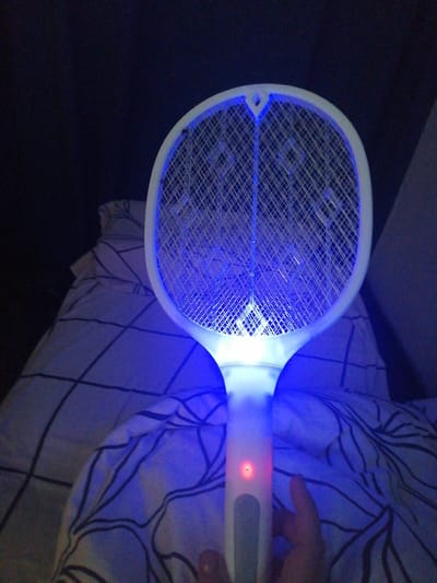 A person's hand is holding a bug zapper that is emitting a bright blue light. The bug zapper is positioned over a bed with patterned sheets. The background is dark, suggesting a nighttime setting. The image appears to have been taken indoors.