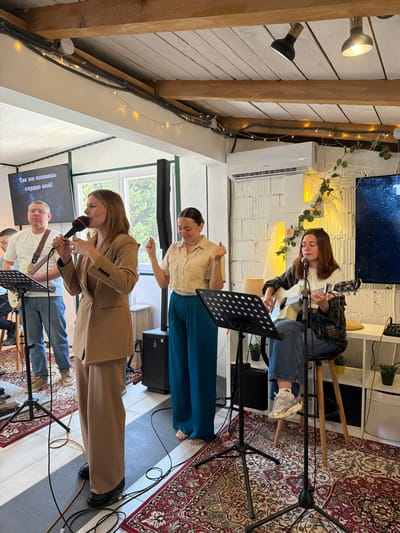 The image depicts an indoor performance with three female vocalists and at least one male guitarist. The location is implied to be in Zgrade, Montenegro. The environment appears to be a repurposed space with exposed wooden beams and whitewashed walls. One vocalist in a tan suit holds a microphone and performs with a male guitarist accompanying her. Above her, another vocalist in a cream blouse and teal pants sings. In the background, a third vocalist plays an acoustic guitar while seated on a stool. Music stands are positioned in front of each performer. A monitor displaying song lyrics is visible behind the lead singer. The floor is covered with a patterned rug. The space is illuminated by various light sources, including overhead spotlights and string lights. A white air conditioner and display screen are mounted on the wall behind the seated performer. The setting suggests a live music event, potentially a recording session or intimate concert.