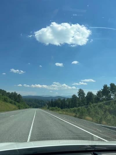 This is a point-of-view photograph taken from inside a vehicle driving on a highway. The road is asphalt with white lane markings, and it stretches towards a distant horizon. On either side of the road, there are green, forested hills and mountains. The sky is bright blue with scattered white clouds. The scene appears to be in a rural or natural setting, with no people or buildings immediately visible. The light conditions suggest it is daytime, likely mid-morning or afternoon. The image does not provide specific details to pinpoint the location within the city of Kaz, Russia, as it depicts a generic highway landscape.