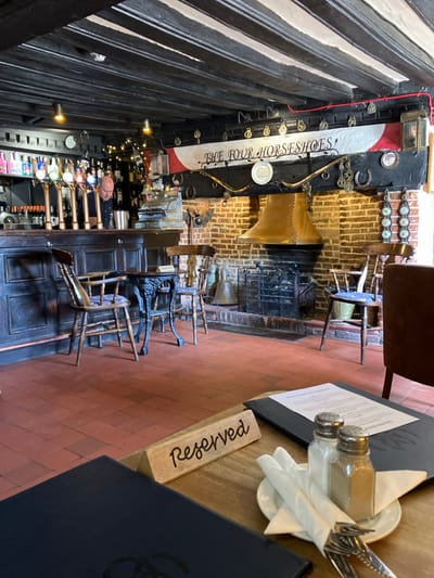The image displays the interior of The Four Horseshoes pub, located in Eye, Mid Suffolk, United Kingdom. The scene is observed from a table in the foreground, looking towards the bar and a large fireplace.
The ceiling features exposed dark wooden beams. The floor is composed of terracotta-colored square tiles.
To the left, a dark wooden bar counter is equipped with at least eight beer taps. Shelves behind the bar are stocked with various bottles. A male individual with short dark hair, wearing a dark shirt, stands behind the bar, looking down into a metallic container. A vintage-style silver cash register is visible on the bar surface to his right.
In the center-right, a prominent brick fireplace occupies the wall, featuring a large, polished copper hood extending over the hearth, which contains a black metal fire grate. Above the fireplace, a banner-like structure displays "THE FOUR HORSESHOES" in script, flanked by red and white sections, and adorned with metallic horseshoe-shaped decorations and other brass objects. A large brass bell rests on the hearth.
Several dark wooden chairs with turned legs and patterned seat cushions are positioned around small tables. One such table, made of ornate black metal, stands between two wooden chairs near the bar. A dark brown upholstered armchair is partially visible in the lower right foreground.
The immediate foreground shows a light-colored wooden table set with two dark menu folders. A wooden, triangular sign displaying "Reserved" in script is placed between them. Adjacent to a menu, a small white plate holds a set of polished cutlery (fork, knife), two white napkins wrapped around the cutlery, and two glass shakers containing salt and pepper.