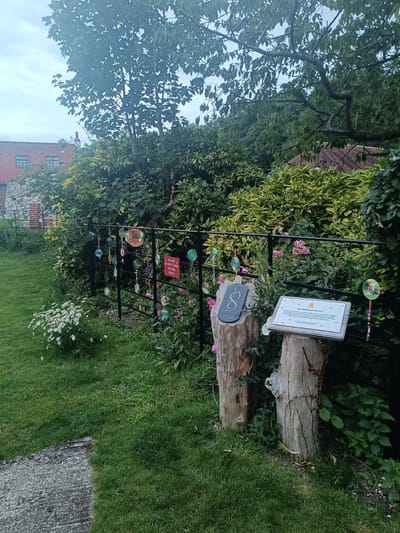 The image shows a garden area with a black metal fence. Several round ornaments are hanging from the fence, some decorated with beads. A red sign on the fence reads "PLEASE DO NOT CLIMB ON THIS FENCE." To the right of the fence, there are two tree stumps. One stump has a stone etched with the number "8" and "2022". A plaque is mounted on a wooden board on the other stump, with the title "The Pollinators Garden" and text that appears to describe the garden. In the background, there is lush green foliage and a brick building. The foreground consists of a grassy lawn and a stone path. The scene appears to be outdoors, with a slightly overcast sky. The location is identified as Arun, United Kingdom. There are no people visible in the image.