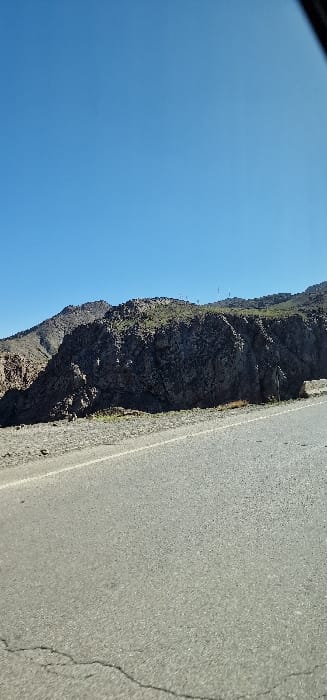 The scene depicts a bright, clear blue sky over a mountainous, arid landscape in Zerkten, Morocco. An asphalt road, showing prominent cracks in the foreground and marked by a solid white line along its right edge, runs horizontally across the lower third of the image. To the right of the road, a narrow dirt shoulder precedes a steep, rocky mountainside characterized by dark grey rock formations and sparse green shrubbery. In the background, additional mountainous terrain is visible, with several slender, vertical structures, consistent with communication towers or antennas, positioned on a distant peak. A small, dark green triangular sign is partially visible at the base of the mountainside adjacent to the road on the far right. No individuals, vehicles, or active elements are present within the frame.