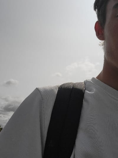 The image shows a person, a young man with short dark hair, wearing a white t-shirt and a black backpack slung over his shoulder. The man is looking to the right of the frame, but the rest of his body is obscured. The sky is overcast with white clouds, and the background is slightly out of focus. The scene is taking place in Calais, France, at the coordinates 50.946, 1.892, most likely near the city center or a park.  The image is taken from the perspective of someone standing behind the man, likely another person. The image is focused on the man and his clothing, with the background blurred, making it appear as though the image is taken in a quiet, introspective moment.  The lack of context or activity further contributes to this impression.