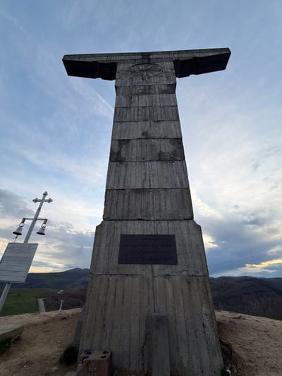 The image depicts the Monument of Georgian Alphabet in Zemo Nichbisi, Georgia. The monument is a large concrete structure with a T-shape at the top and a relief of an ancient Georgian symbol on the front. A plaque with text is affixed to the side of the monument. The surrounding landscape includes mountainous terrain and a cloudy sky. In the foreground, there is a cross-shaped bell tower and a wooden sign. The monument stands on a cliff edge with vegetation nearby.