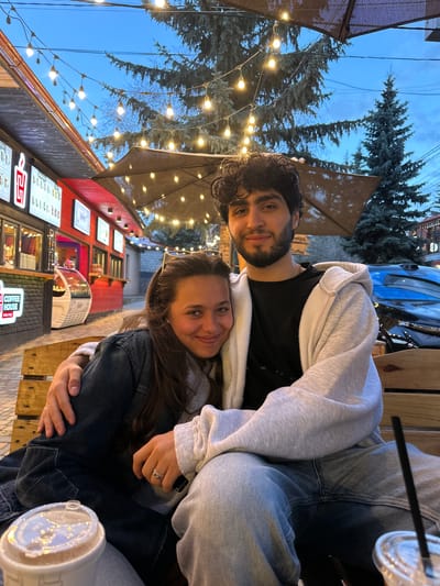 A couple is seated close together at an outdoor cafe in Tsakhkadzor, Armenia. The man has dark curly hair and a beard, wearing a black shirt and a light grey hoodie. The woman has long brown hair and is wearing a denim jacket. They are situated at a wooden table with drinks; a plastic cup with a lid and straw is visible. Behind them is a red building with illuminated menu boards and cafe signage indicating "Coffee House" in both English and Chinese. String lights illuminate the area and tall evergreen trees are visible, with a glimpse of a blue car and twilight sky.