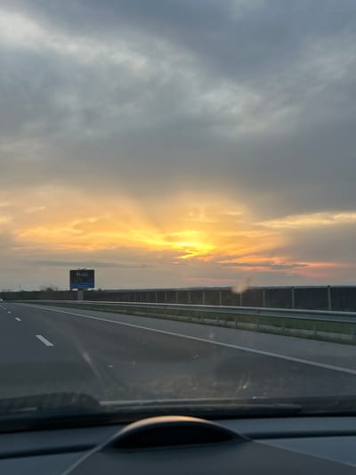 The image shows a view from inside a vehicle traveling on a highway. The sky is predominantly cloudy with the sun setting or rising, casting orange and yellow light through the clouds. A highway sign is visible next to the road. The road surface is grey asphalt with white lane markings and guardrails are seen beside the road. The image appears to be taken from the passenger seat. There is no other information that indicates the location is in the city of Cherna Gora, Bulgaria.