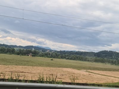 The image shows a rural landscape under a cloudy sky. In the foreground, there is a field with dry, cut vegetation, possibly straw or hay. Beyond this field, a green, mowed field stretches towards a wooded area. In the distance, a village with houses and church steeples is visible nestled among rolling hills and dense forests. Mountains form the backdrop, and power lines are strung across the sky. The scene appears to be in the vicinity of Hörzendorf, Austria, given the characteristic topography and architecture. There are no people or animals visible in the image. The overall impression is one of a tranquil, agricultural setting.