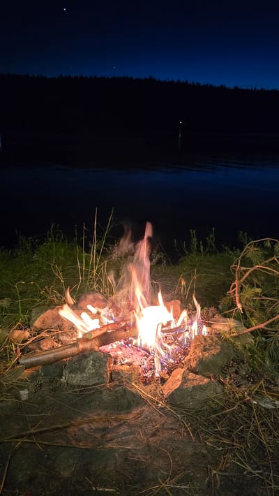 An outdoor campfire is burning at night, situated on the shore of a body of water. The flames are bright and intense, casting a warm glow on the surrounding rocks and dry vegetation. The background is dark, with a dense line of trees silhouetted against the night sky. The sky itself is a deep blue, with a faint crescent moon visible and a distant, small light reflecting on the water. The scene evokes a sense of wilderness and solitude. There are no people present.