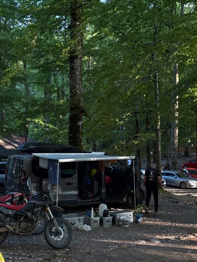 In Sjerogošte, Montenegro, a campsite is set up in a forest clearing. A grey van, outfitted with a roof rack and an awning, is parked on the gravelly ground. Two motorcycles, one red and black and the other black and red, are positioned in front of the van. Various camping gear, including metal containers, a bucket, and a water jug, are arranged on the ground near the van. A person wearing a black t-shirt and black pants is walking away from the van towards a silver sedan and red cars parked in the background. The scene is surrounded by tall trees with dense green foliage, indicating a natural, wooded environment.