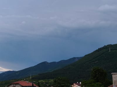 This image presents a serene mountainous landscape under a heavily overcast sky, likely depicting an area in Krichim, Bulgaria. Dominant in the frame are rolling mountains, blanketed with dense, dark green forests, extending from the middle ground into the hazy distance. Distinct ridges and some rocky outcrops are visible along the mountain contours, especially on the left side.

The sky occupies the upper portion of the image, filled with uniform, grey-blue clouds that suggest a cloudy day, possibly indicating impending rain or recent stormy weather. The light is diffused and soft, creating a subdued and somewhat dramatic atmosphere.

In the foreground and middle ground, several residential buildings are partially visible, hinting at a small village or rural settlement. On the left, a house with a prominent terracotta-tiled roof and light-colored walls stands out, nestled among trees. Another similar building is partially seen on the far right. Abundant green foliage and trees are scattered across the lower parts of the frame and climb the mountain slopes.

Man-made elements include two slender lattice towers, possibly for electricity transmission or communication, which are perched on the forested mountain slopes; one is clearly visible on the right slope, and another smaller one appears further left in the mid-ground.

The overall impression is a tranquil, rural setting amidst impressive natural scenery, captured under the muted light of an overcast day, likely in the late afternoon or early evening. There are no people visible, and no specific activities are taking place.