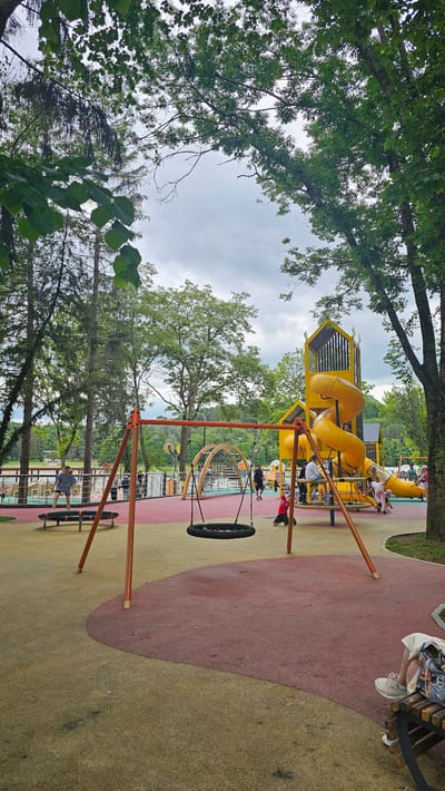 The image depicts a vibrant outdoor playground located in Alikonovka, Russia. The playground features a large yellow multi-level structure with slides and climbing elements, a prominent orange swing set with a black tire swing, and a spinning disc play structure. The ground is covered with a soft, shock-absorbent material in contrasting colors of red and yellow. Several people, including children and adults, are visible enjoying the playground. In the background, tall trees frame the scene, and a body of water, possibly a lake or river, can be seen beyond the playground. The sky is overcast with clouds, suggesting a mild or slightly humid day. The overall impression is of a well-maintained and active recreational space within a natural setting.